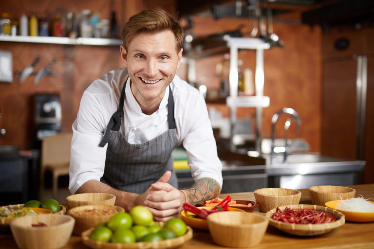 Portrait smiling chef leaning on counter with fruit and veg.