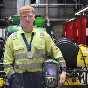 Leaner Luke wearing medal and holding welding helmet in workshop.